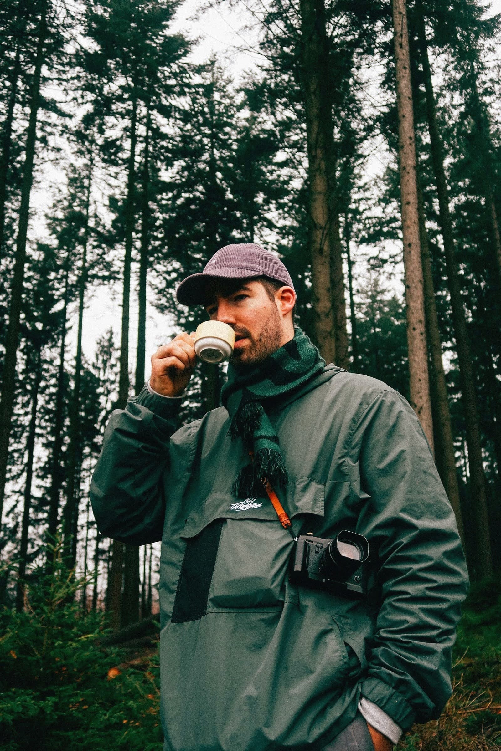 Jonas Denil drinking coffee in a pine forest with a camera around his neck
